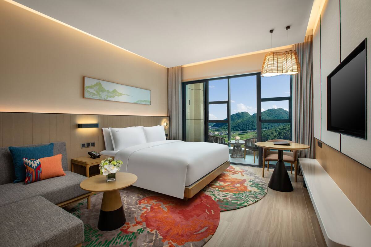 A modern hotel room with a large bed, a sofa, a small table, a TV, and a balcony with a mountain view.
