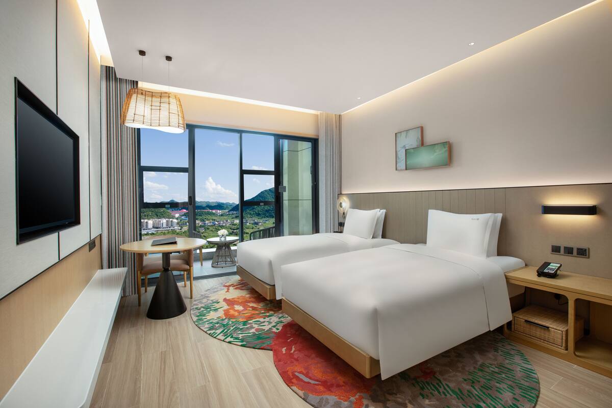 A hotel room with two beds, a TV, a small table, and a view of the city.
