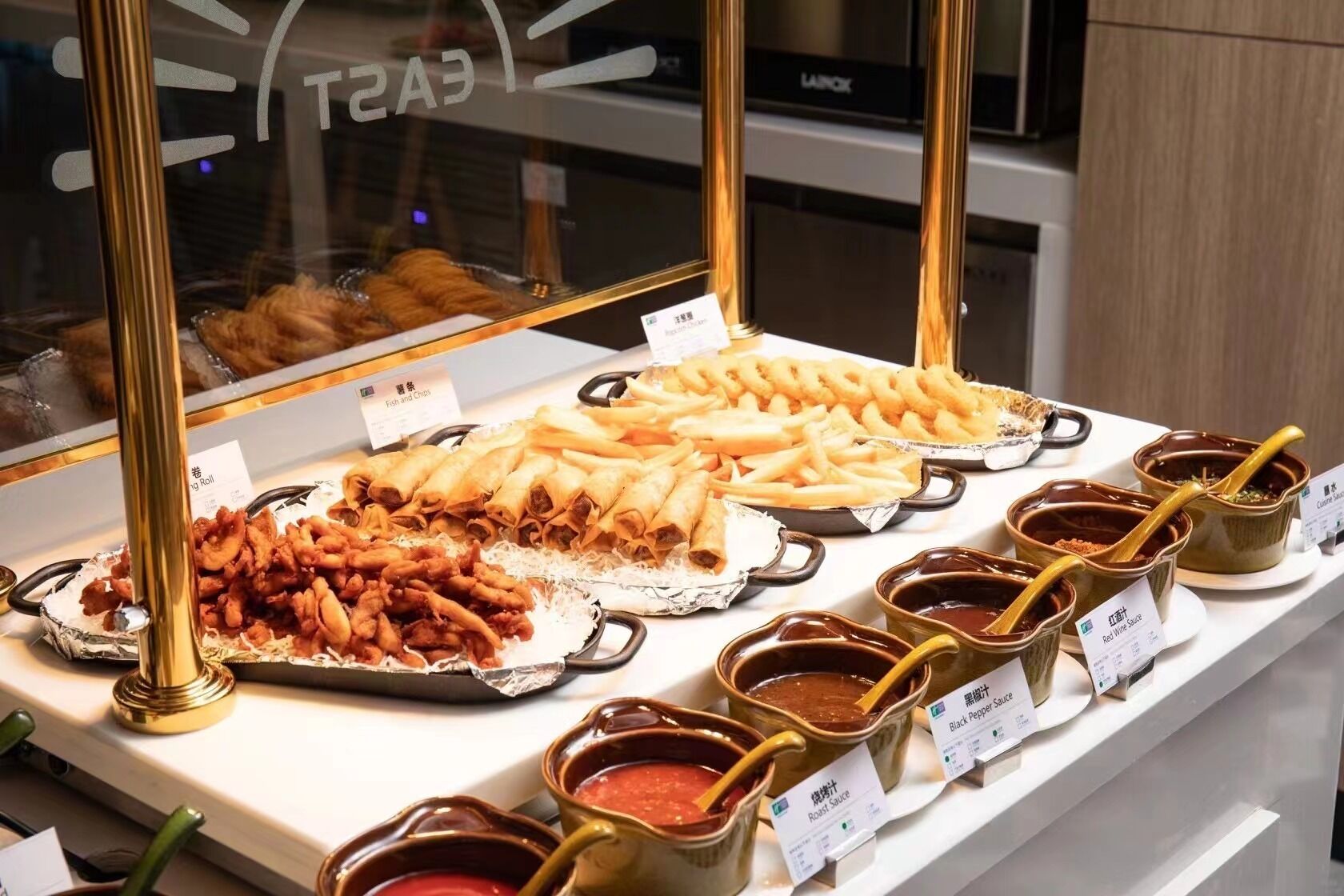 A buffet with various dishes and sauces, including fried items and rolls, displayed in a glass case.