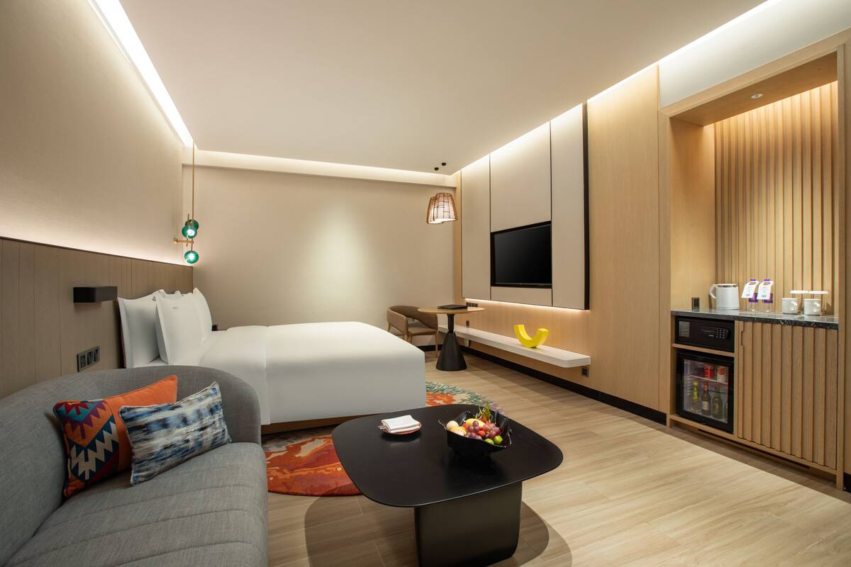 A modern hotel room with a bed, a sofa, a coffee table with fruit, a TV, a small table, and a minibar.