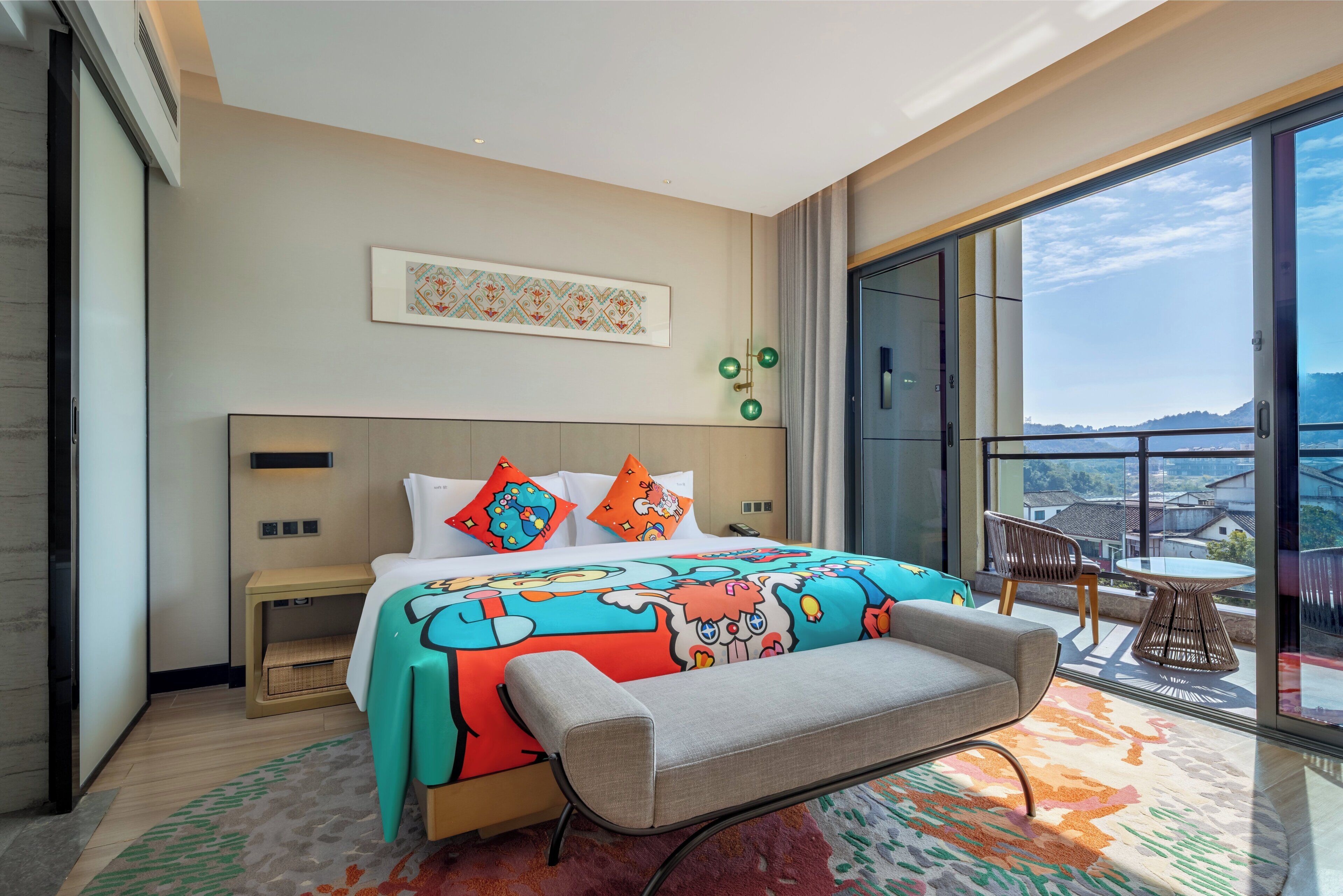 A modern hotel room with a large bed, colorful bedding, a bench, a small table, and a balcony with a view.