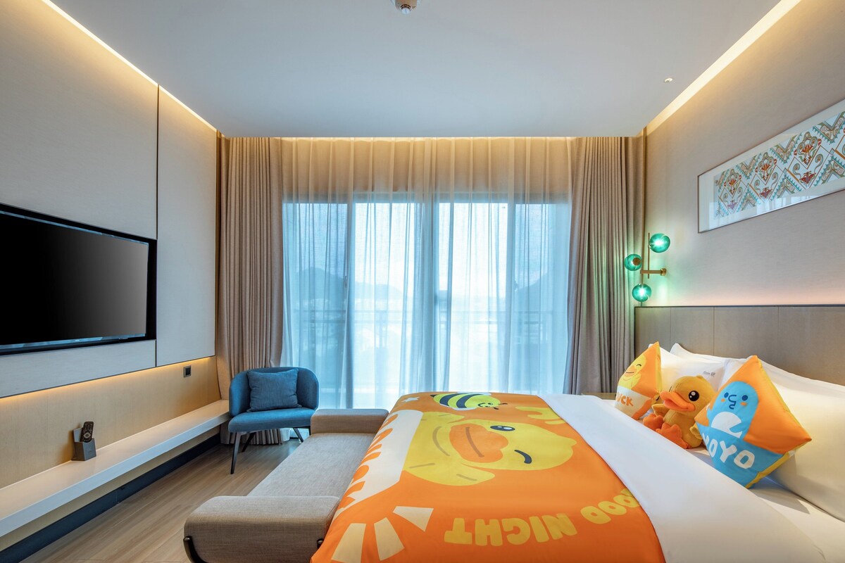 A hotel room with a large bed, a TV mounted on the wall, a seating area with a chair, and a window with curtains.