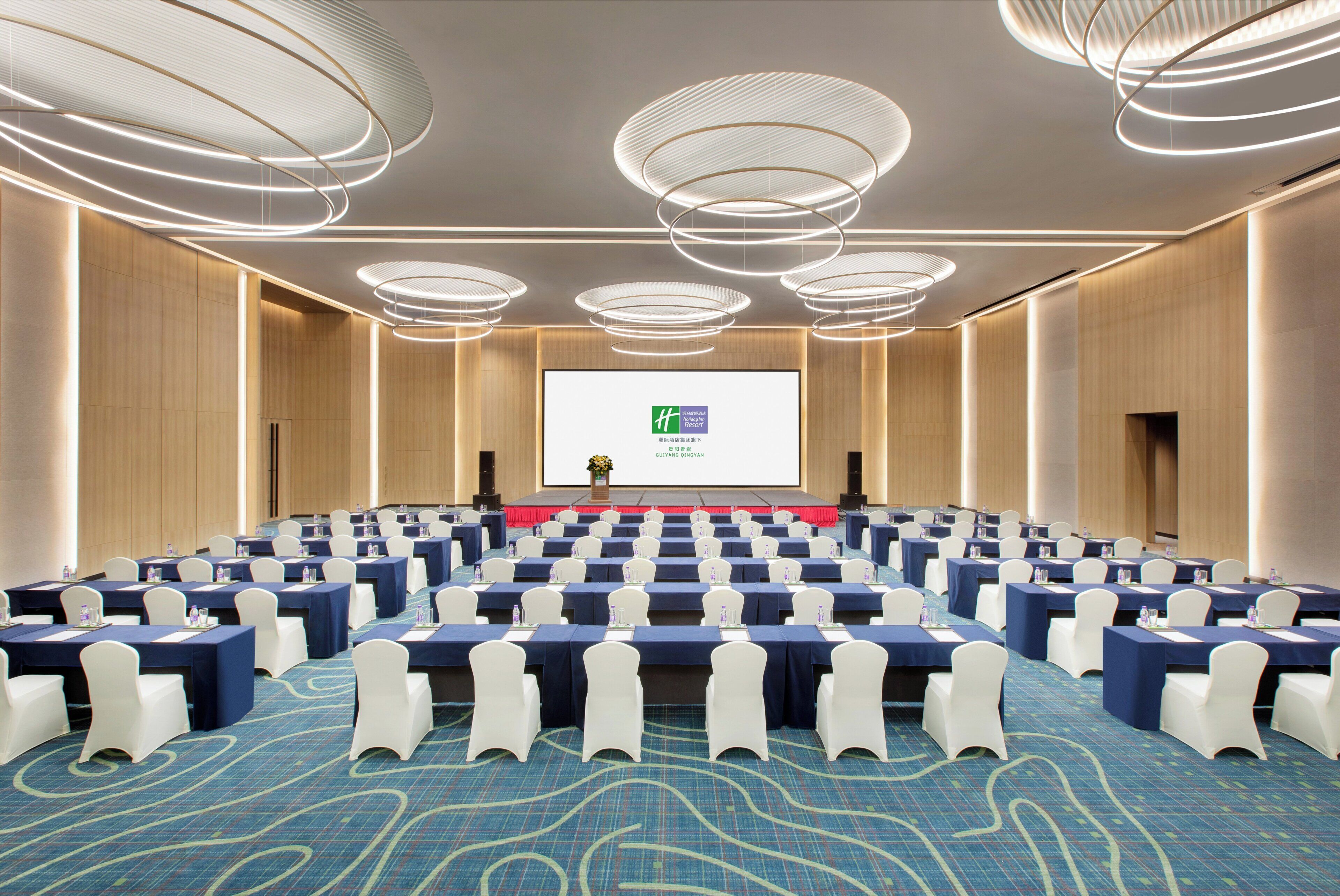 Un salón de conferencias con filas de mesas, sillas blancas y una pantalla grande al frente.