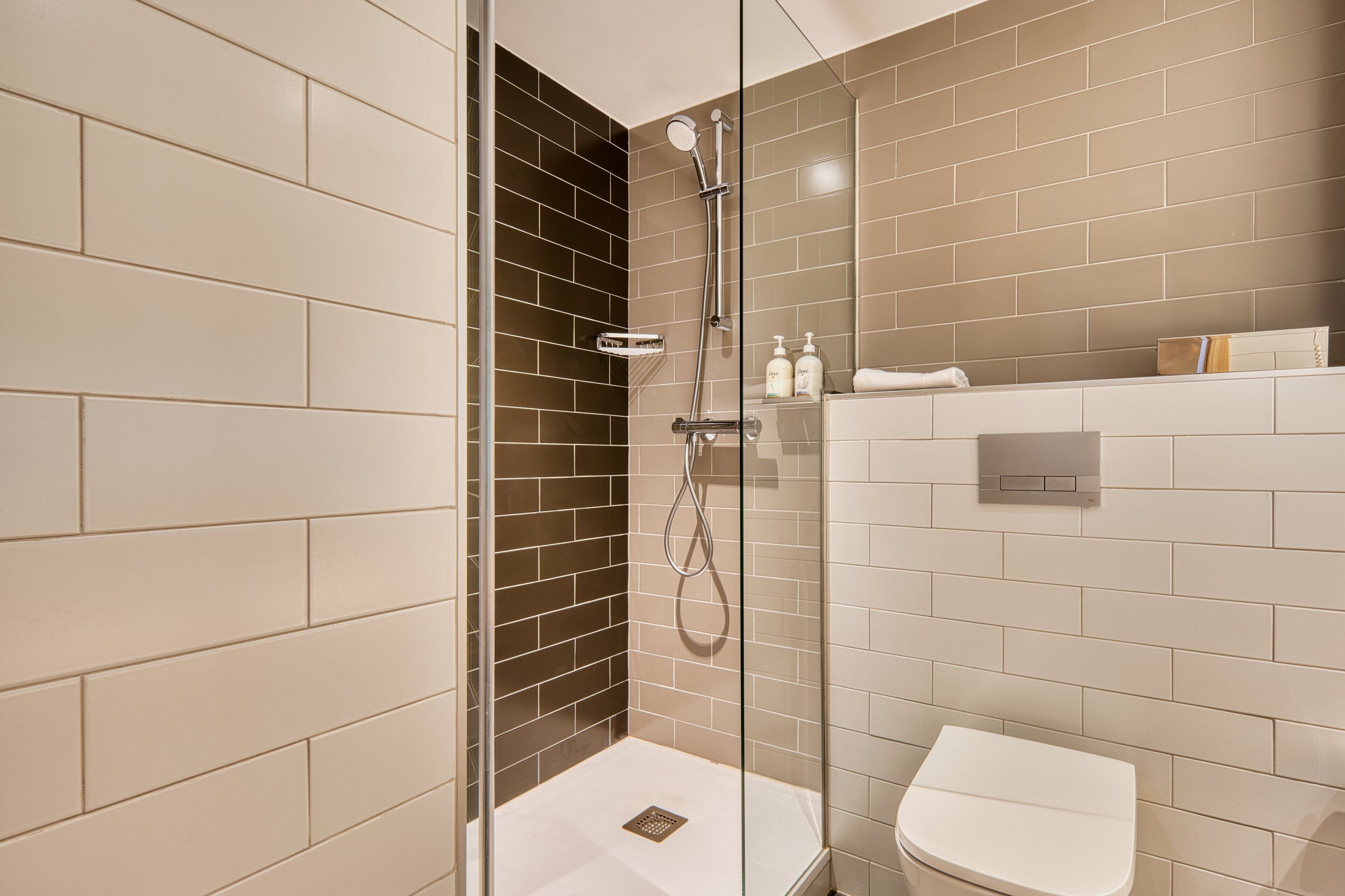 A modern bathroom with a glass-enclosed shower, a white toilet, and a tiled wall with a mix of beige and dark brown tiles.