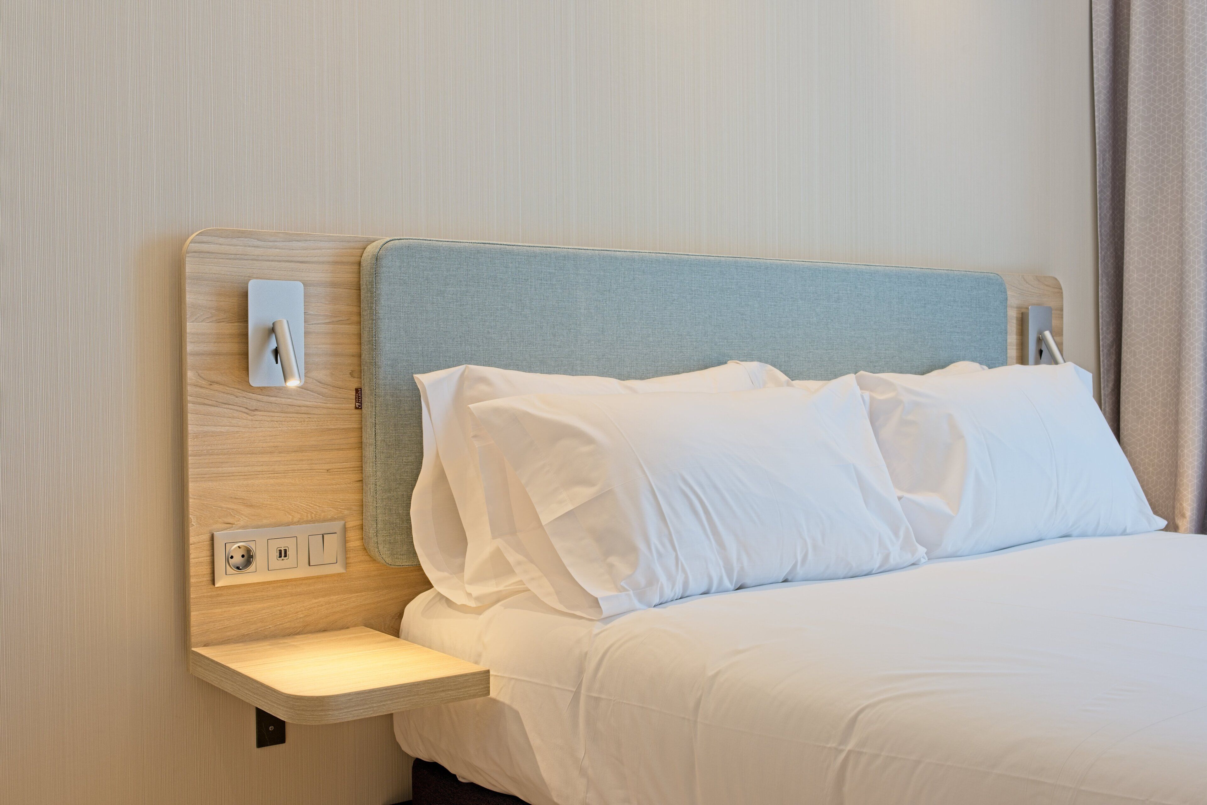 A neatly made bed with white linens, a wooden nightstand with a light switch, and a wall-mounted outlet.
