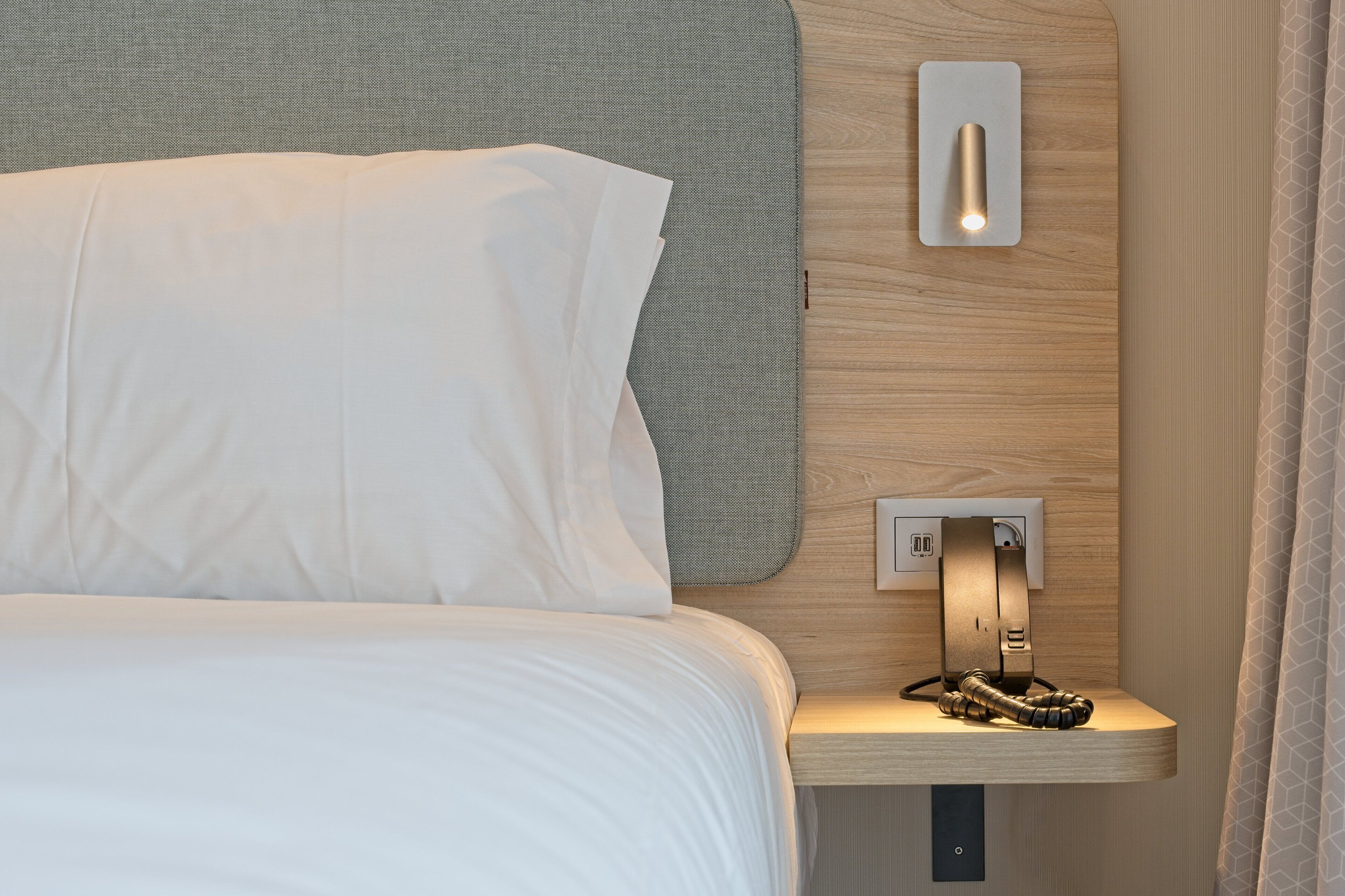 Un lit bien fait, avec du linge de lit blanc, une table de chevet avec une lampe et une prise de courant.