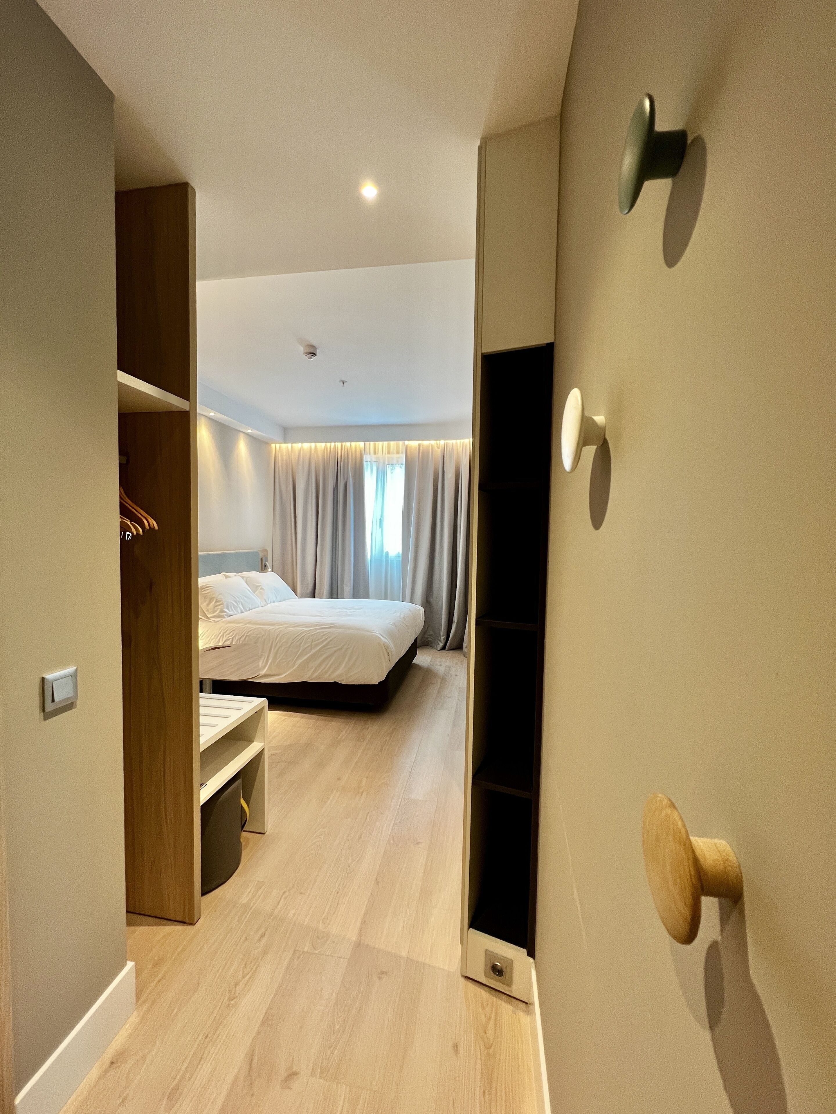 A modern hotel room with a wooden wardrobe, a bed with white bedding, a small desk, and a built-in TV.