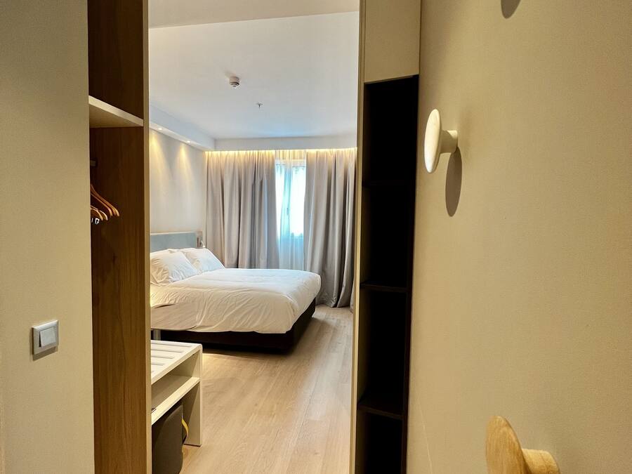 Una habitación de hotel moderna con un armario de madera, una cama con ropa blanca, un escritorio pequeño y un televisor integrado.