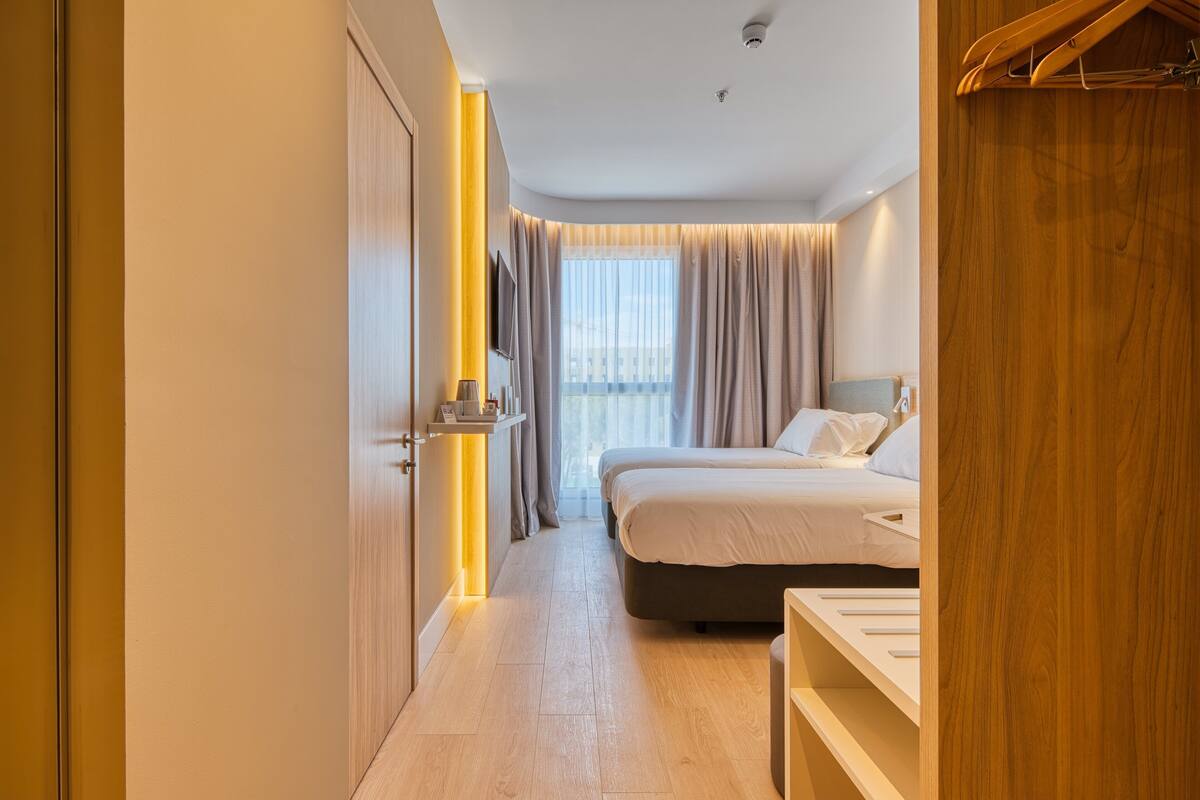 A modern hotel room with a large bed, bedside tables, a TV, and a view of the city through large windows.