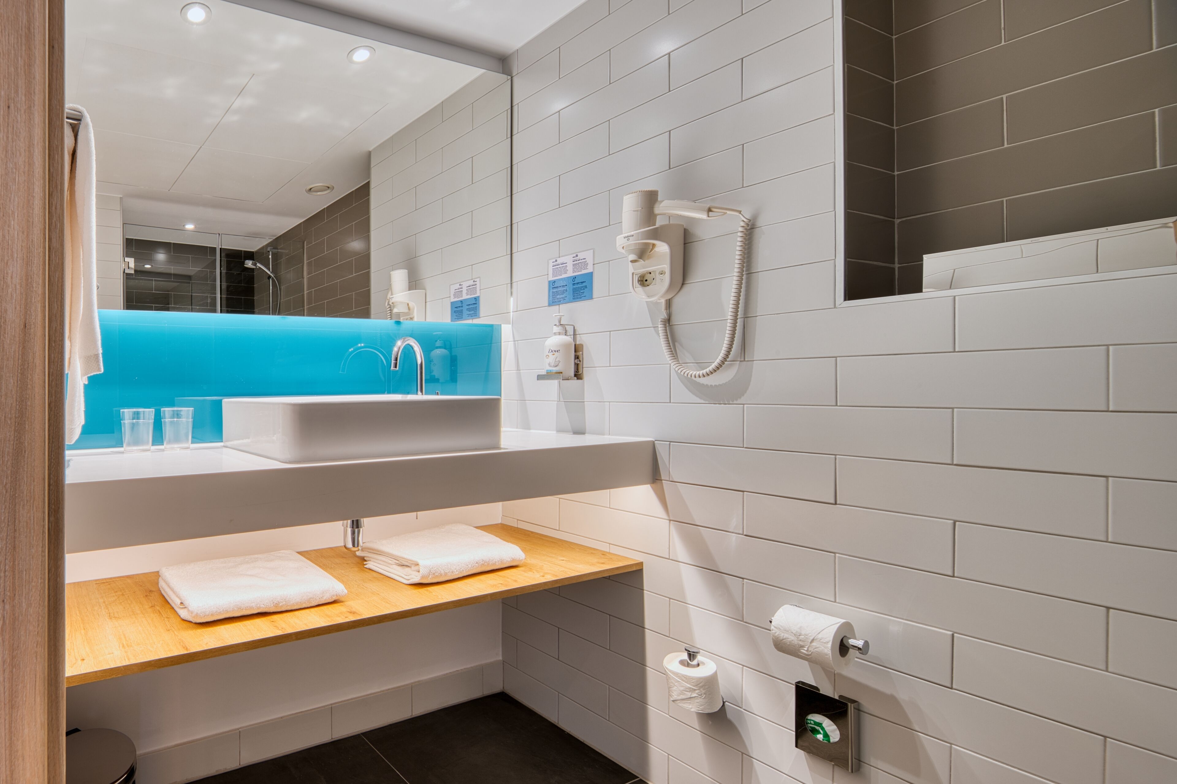A modern bathroom with a blue sink, white tiles, a wooden shelf with towels, a mirror, and a hand dryer.