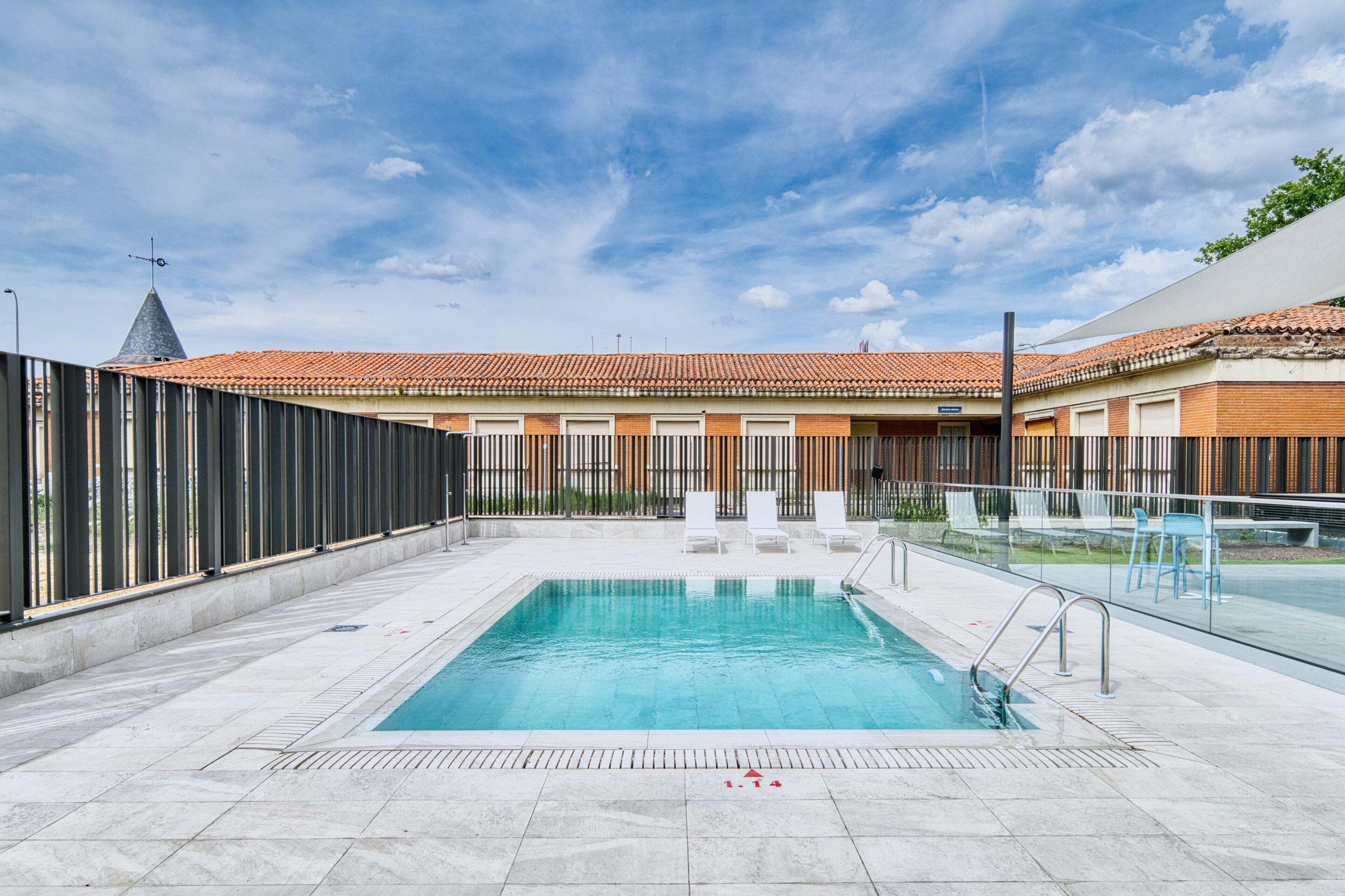 Una piscina con borde de baldosas, un edificio de ladrillo con tejado de tejas rojas y una cerca de metal.