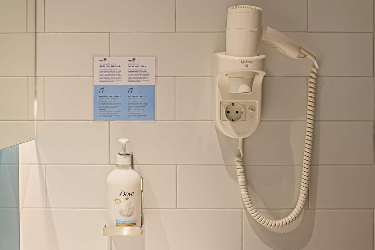 A bathroom with a hairdryer mounted on the wall, a hand sanitizer dispenser, and a sign with water conservation tips.