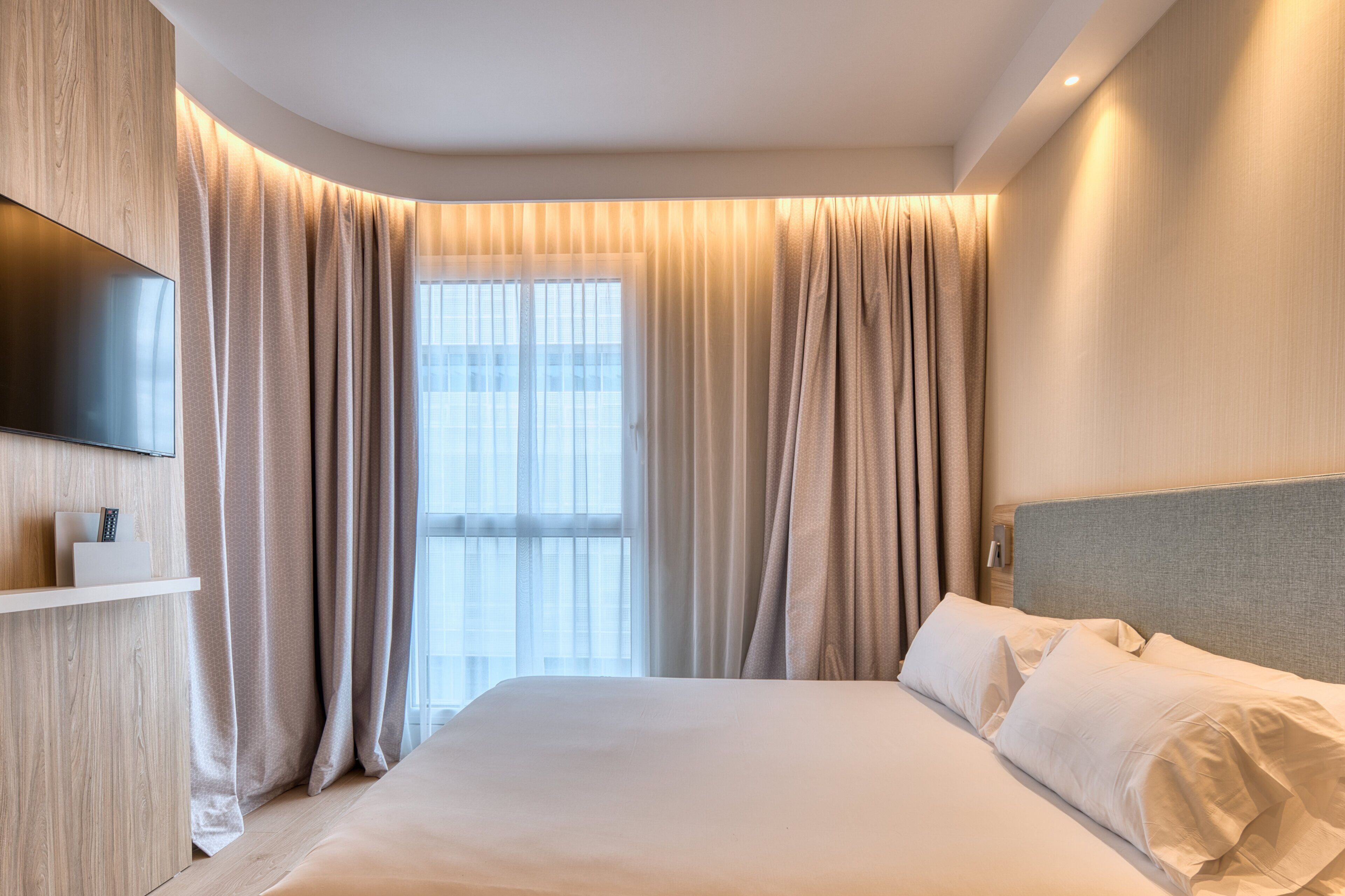 Une chambre d’hôtel moderne dotée d’un grand lit, d’un téléviseur à écran plat fixé au mur et d’une grande fenêtre avec des rideaux.