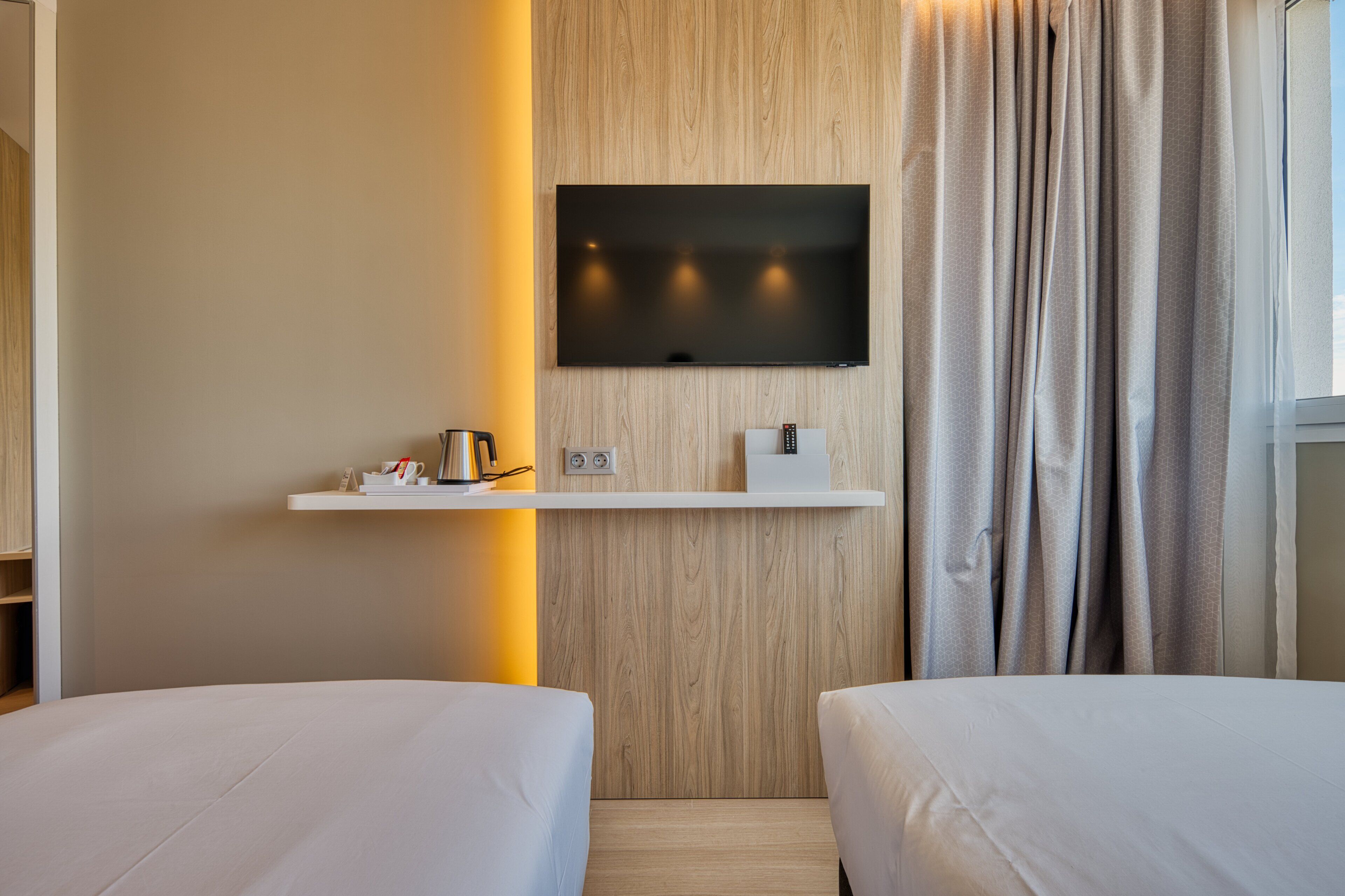 Ein Hotelzimmer mit zwei Betten, einem Wand-Fernseher, einem Nachttisch mit Lampe und einem Fenster mit Vorhängen.
