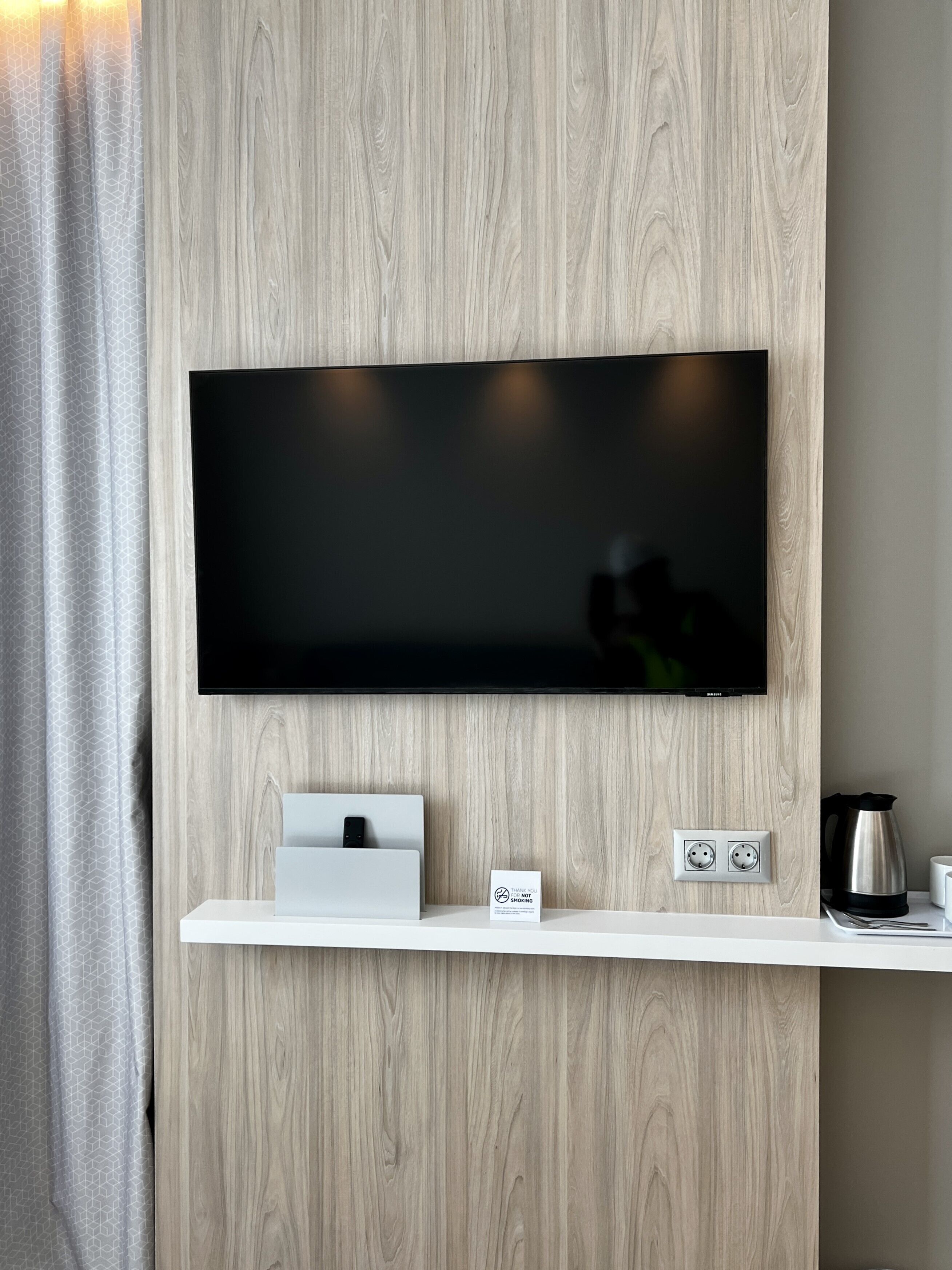 Una habitación moderna con un televisor de pantalla plana montado, una estación de carga blanca y un pequeño estante con una tetera y tazas.