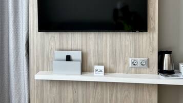 Una habitación moderna con un televisor de pantalla plana montado, una estación de carga blanca y un pequeño estante con una tetera y tazas.