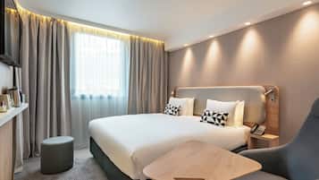 A modern hotel room with a large bed, a bedside table, a chair, a small stool, and a TV.