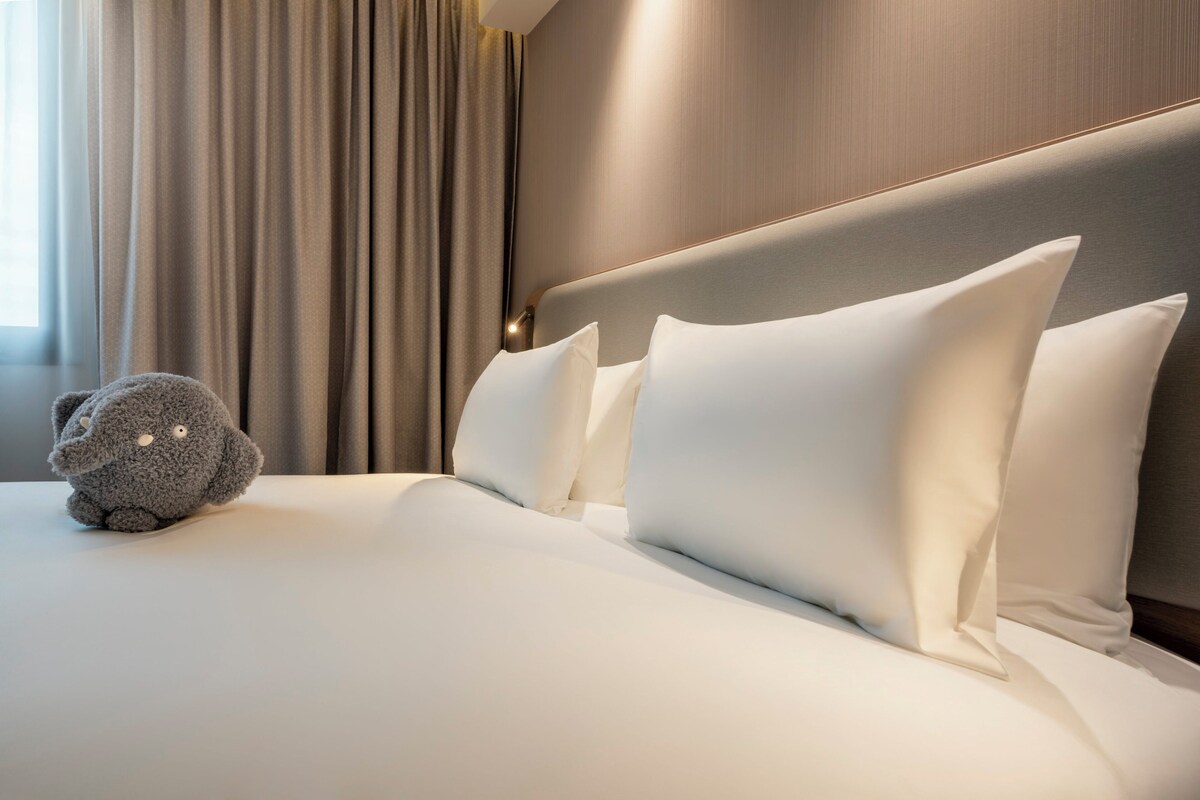 Una cama bien hecha con sábanas y almohadas blancas, y un peluche sobre la cama, junto con un cabecero moderno.