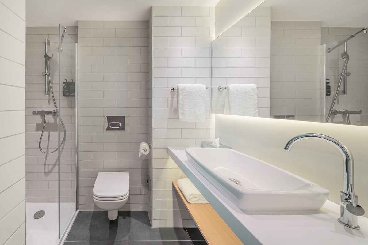 Un baño moderno con ducha de vidrio, inodoro blanco, un lavabo grande con espejo y un perchero para toallas.