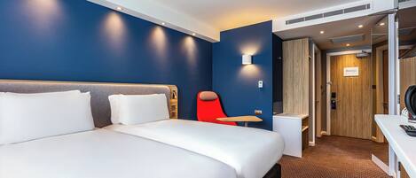 A hotel room with a large bed, a small desk, a red chair, and a wooden door.
