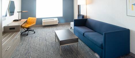 Una habitación de hotel moderna con un sofá azul, un escritorio de madera, una silla naranja, una mesita pequeña, una lámpara de pie y un ventanal con persianas.