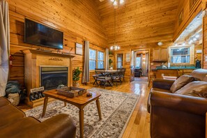 A log cabin interior with a fireplace, a television mounted above it, a dining area with a table and chairs, and a living room with a sofa and a coffee table.