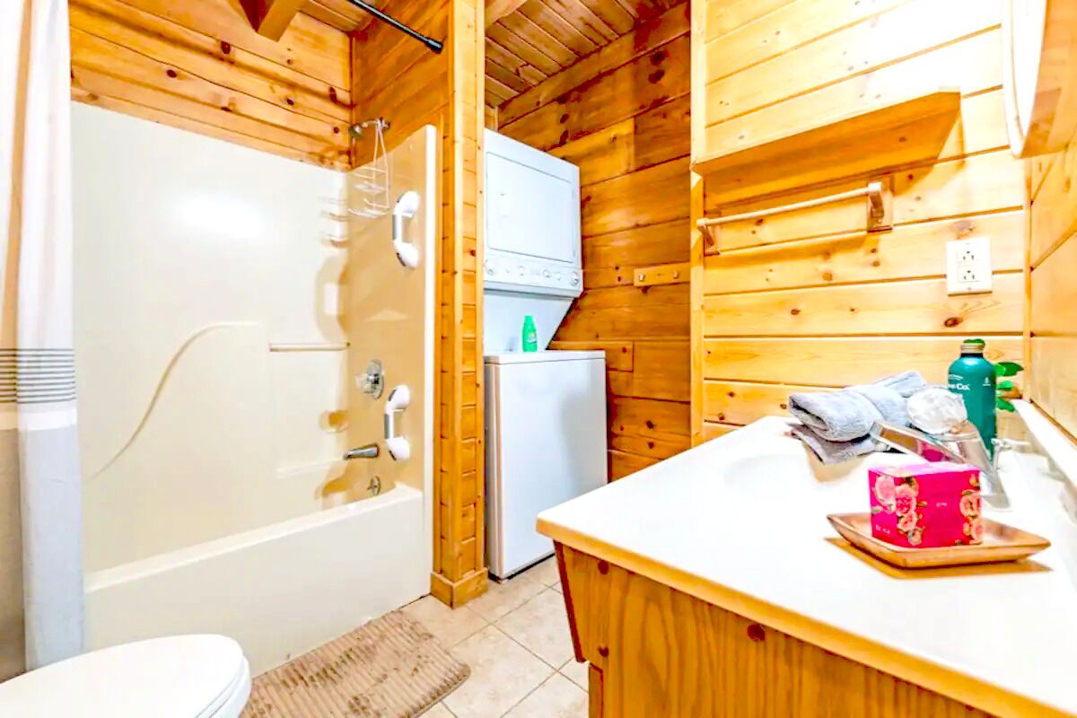 A wooden cabin bathroom with a bathtub, a white toilet, a sink with a mirror, and a refrigerator.