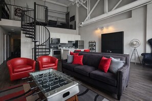A modern living room with a sofa, red armchairs, a foosball table, a staircase, a kitchen, and a black TV.