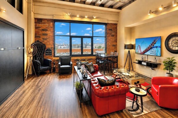 A modern living room with a large window, a red sofa, a dining area, and a television displaying Cedar Point. - Downtown Loft By Cedar Point Sports Force! (Sandusky)
