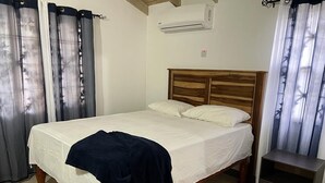 A bedroom with a wooden bed, white bedding, a dark blue throw blanket, a nightstand, and a wall-mounted air conditioner. - Vacation Home Rental (Florence Hall Village)