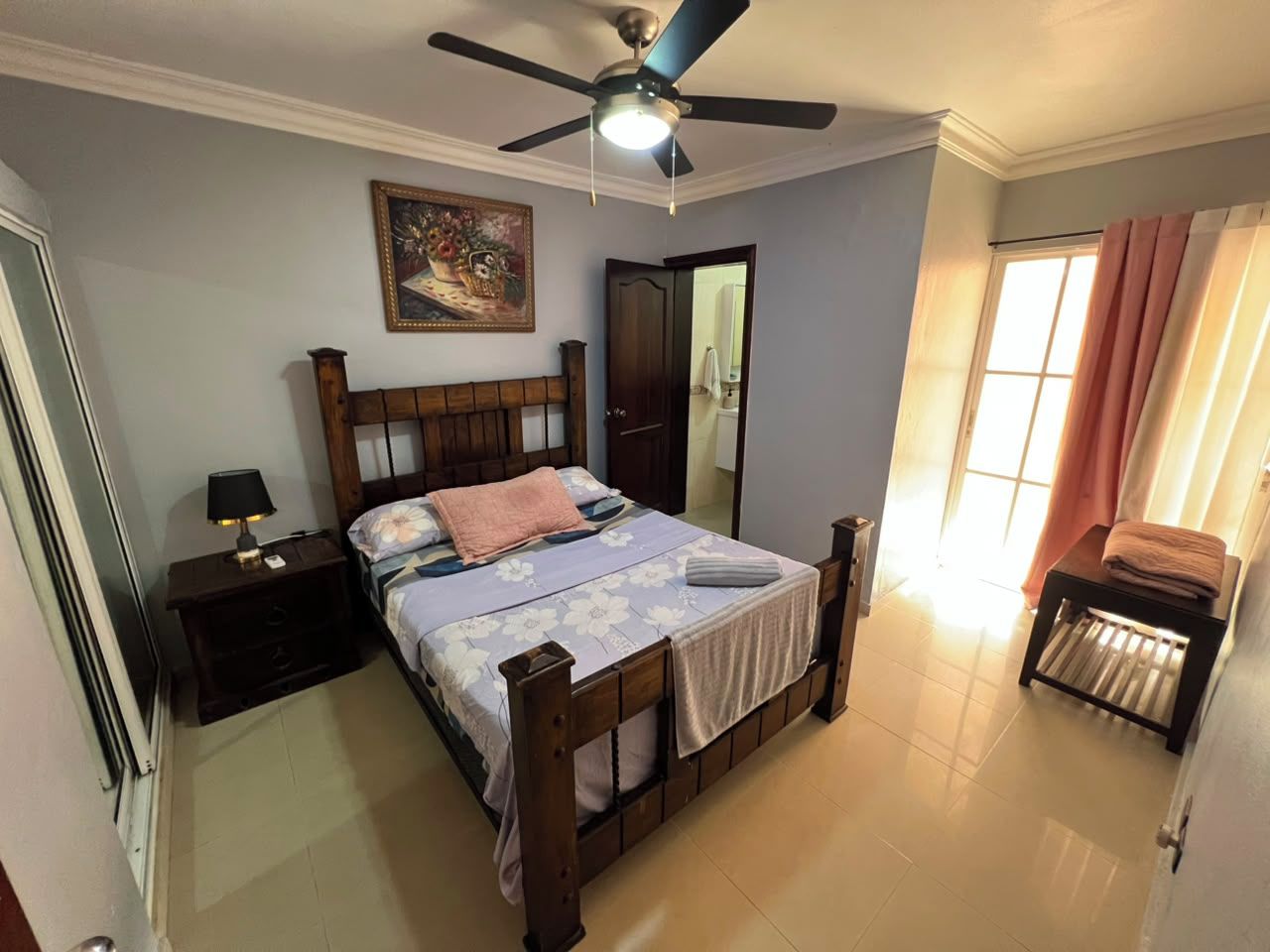 A bedroom with a bed, a nightstand, a ceiling fan, a door, and a window with curtains.