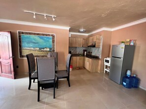A dining area with a table and chairs, a kitchen with wooden cabinets, a refrigerator, and a water dispenser.