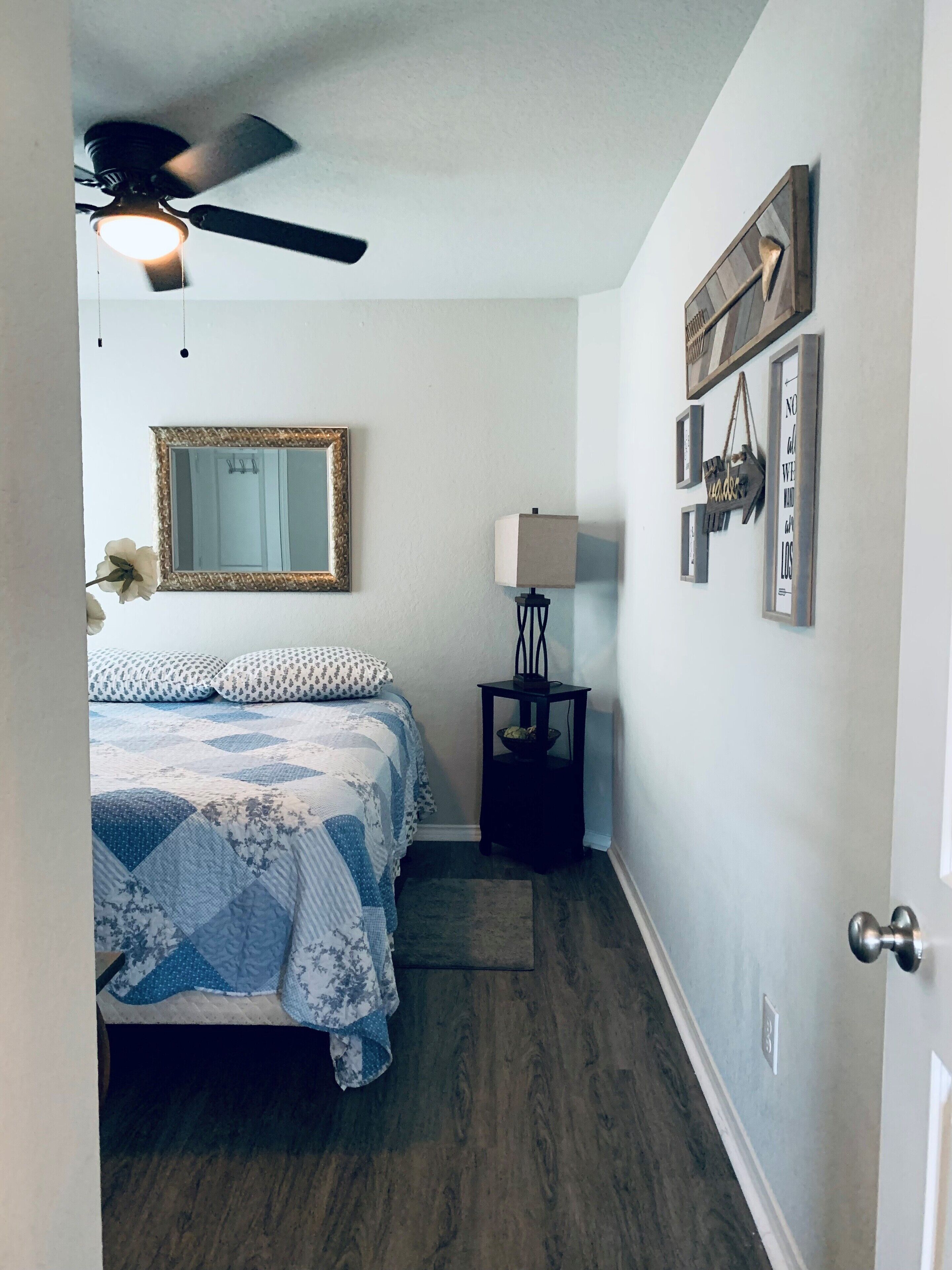 A bedroom with a bed, a ceiling fan, a mirror, a nightstand with a lamp, and wall decorations.