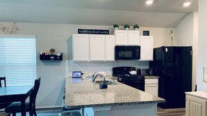 Una cocina moderna con armarios blancos, un refrigerador negro, un microondas y una estufa. Hay una zona de comedor con una mesa y sillas.