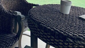 Un patio techado con sillas tejidas y una mesa, una taza de cerámica blanca, y vista a un jardín con césped, una valla de madera y árboles.