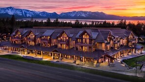 A large, multi-story lodge with a symmetrical roofline and multiple balconies, set against a backdrop of mountains and a lake at sunset.
