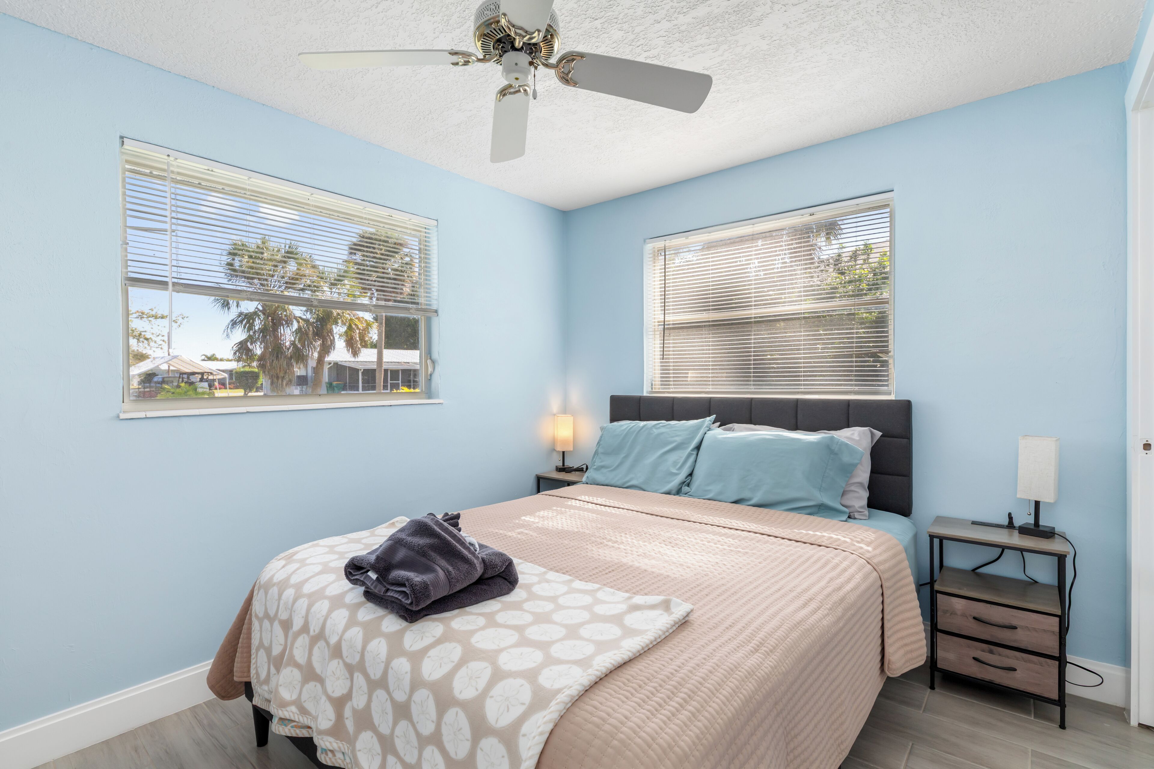 A bedroom with a bed, two bedside tables, a ceiling fan, and windows with blinds.
