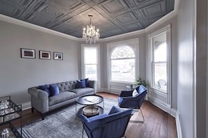 Un salon avec un canapé gris, des fauteuils bleus, une table basse ronde et un lustre. Trois tableaux encadrés ornent le mur, et de grandes fenêtres sont agrémentées de motifs décoratifs en fer forgé.