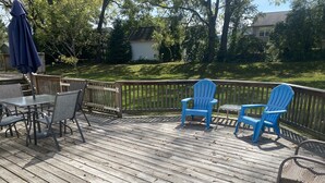 A wooden deck with a table and chairs, a blue umbrella, and a view of a house and trees. - Our Comfortable Neighborhood Home (Boalsburg)