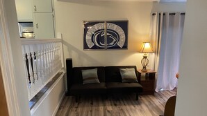 A living room with a dark sofa, two cushions, a wall-mounted sign, a lamp, and a wooden floor. - Our Comfortable Neighborhood Home (Boalsburg)