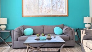 Ein wohnzimmer mit tealfarbenen Wänden, einem grauen Sofa, zwei tealfarbenen Kissen, einem Couchtisch mit einem Schild und einem raumhohen Fenster.