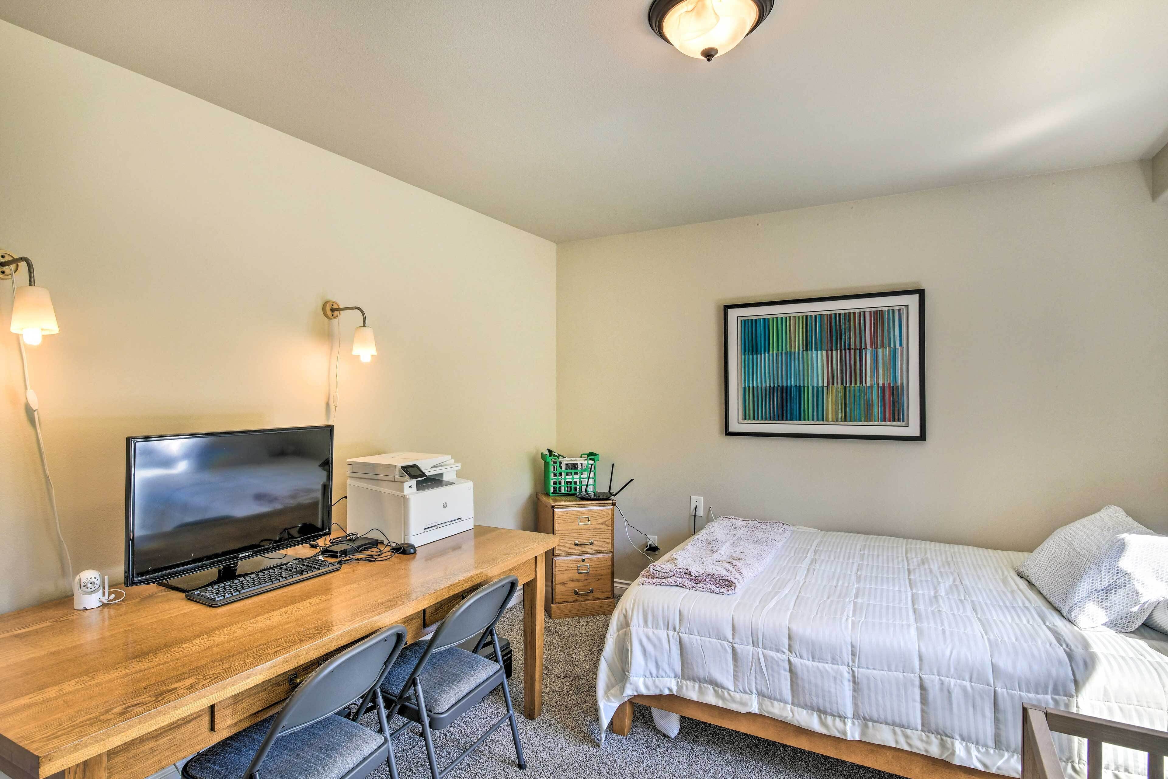 A bedroom with a wooden desk, a bed with a white comforter, a wall-mounted abstract painting, and a ceiling light fixture.