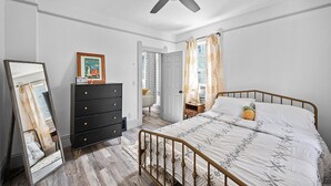 Una camera da letto con un letto, una cassettiera, uno specchio, una ventola a soffitto e una porta che dà su un'altra stanza.