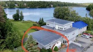 Une maison à deux étages avec un toit bleu, entourée de verdure et bordée d’un lac.