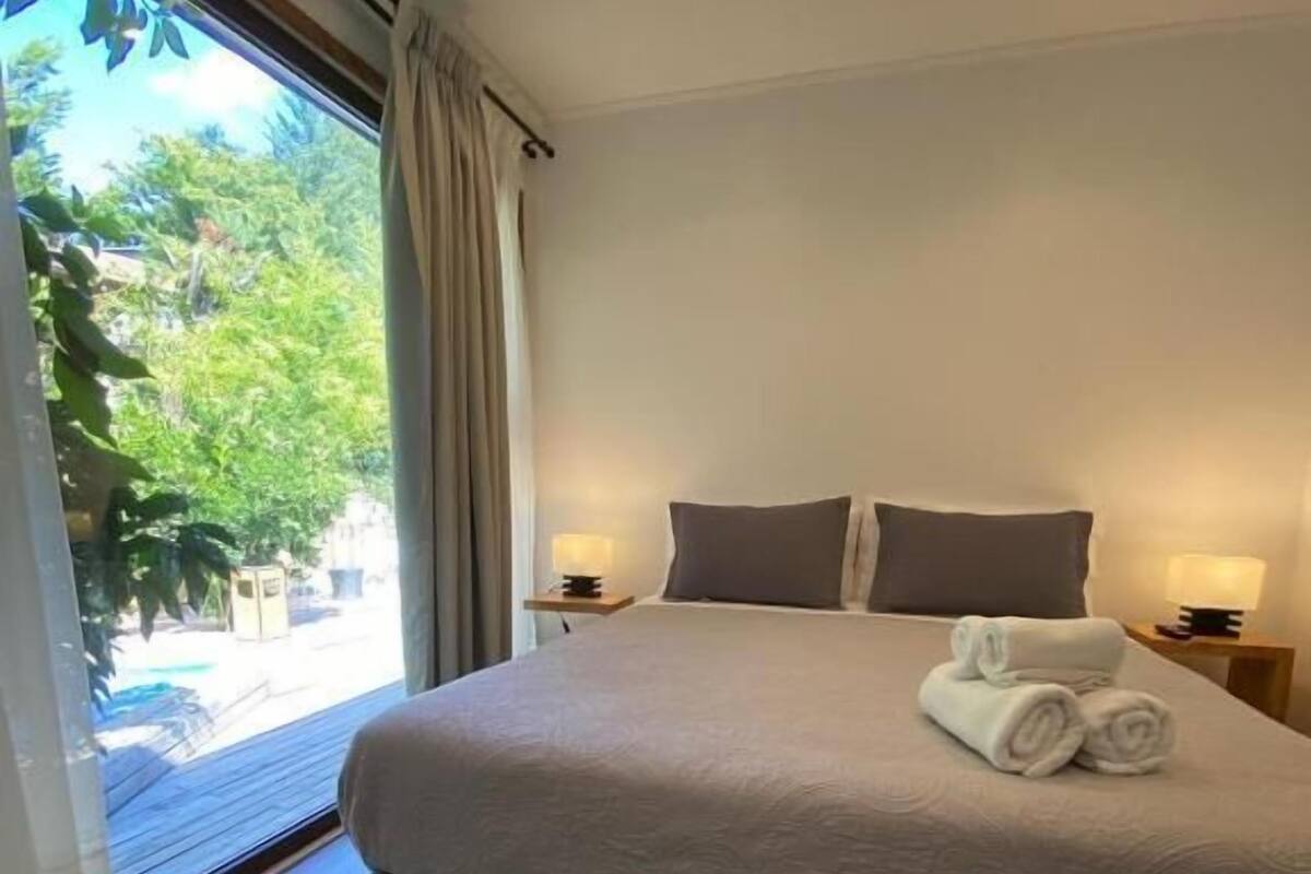 A large bed with a neutral-colored bedspread and two pillows, a small bedside table with a lamp, and a view of greenery outside.