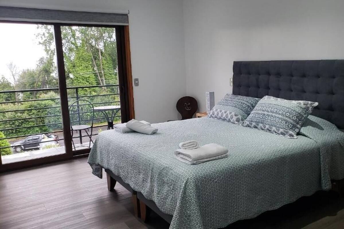 A bedroom with a large bed, a balcony with a table and chairs, and a view of greenery outside.