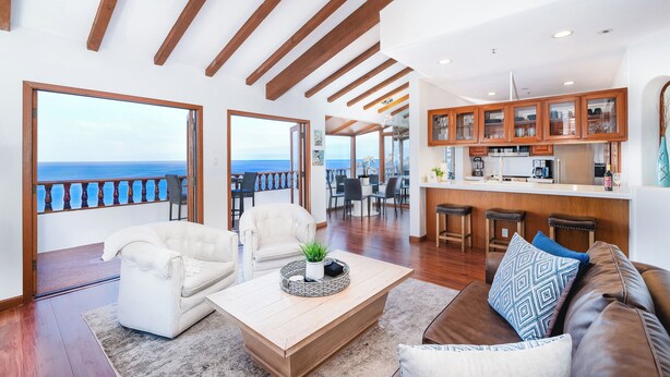 Amplia sala de estar con vista al mar, vigas de madera en el techo y una cocina moderna con taburetes de bar.