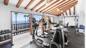 Un gimnasio con cintas de correr, elípticas y pesas libres, con vistas a un balcón frente al océano.