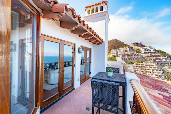 A balcony with a view of a coastal town, featuring a table and chairs, and a door leading to an outdoor seating area.