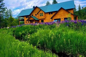 A log cabin with a metal roof, surrounded by lush greenery and purple flowers.